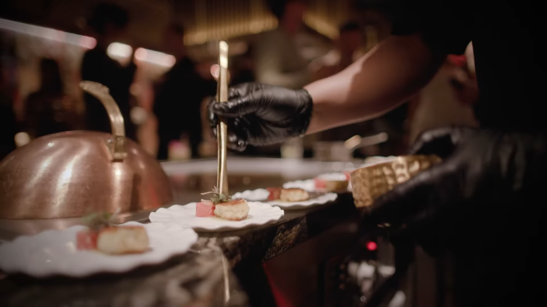 A Chef in Black Gloves Carefully Plates a Gourmet Dish with Precision at A Restaurant in Downtown La, with A Busy Dining Atmosphere in The Background